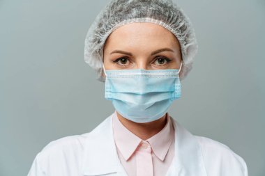 Gri arka planda tıbbi maske takan mutsuz genç bir doktorun portresi. Doktorlar yorgun gözlerle baktı. Koronavirüs konsepti.