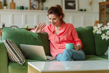 Portrait of a woman sitting on the couch at home, communicating via video link with relatives. Happy young woman talking on webcam, online course indoors.