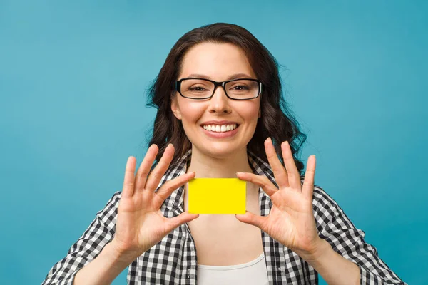 Mulher sorridente bonito em camisa de vestido e óculos, mostrando cartão de crédito na mão para o conceito da sociedade financeira e sem dinheiro, posando em fundo azul. Espaço de cópia. — Fotografia de Stock