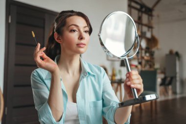 Genç ve güzel bir kadın aynaya bakarken evde makyaj yapıyor.