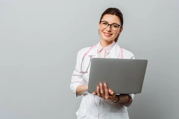 Portret van een vriendelijke lachende jonge vrouwelijke arts. Afbeelding van een vrouwelijke arts met bril en laptop. Geïsoleerd op een grijze achtergrond. — Stockfoto