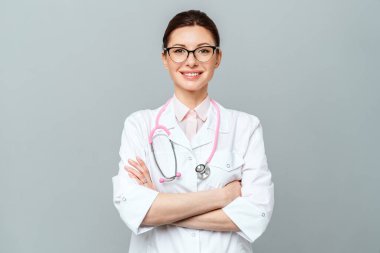 Beyaz üniformalı gülümseyen doktorun portresi gri arka planda çapraz ellerle duruyor.