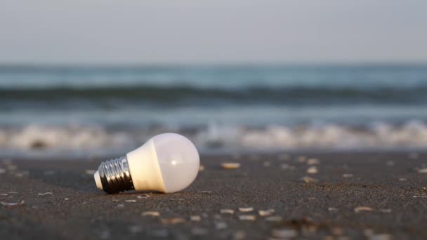 Used led light bulb lies on wet sand of beach against waves — Video Stock