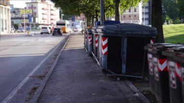Siyah plastik çöp tenekeleri kaldırımda arabalara karşı duruyor.