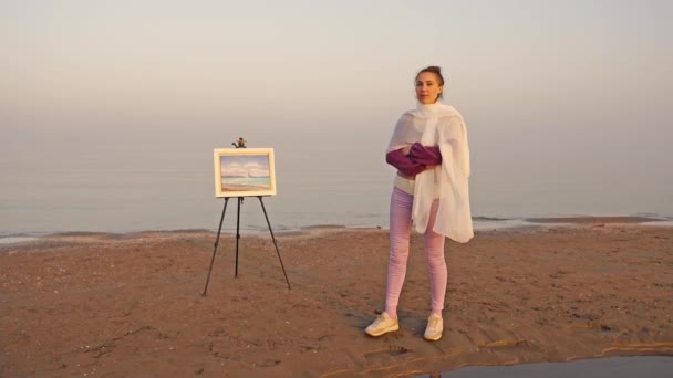 Frau mit weißem Schal steht am Strand an Staffelei mit Meerblick — Stockvideo