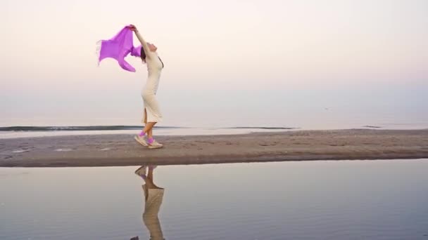 Wanita dengan gaun putih berjalan di pantai melambaikan syal ungu — Stok Video