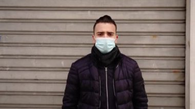 Koruyucu maskeli adam kapalı mağazanın kepenklerinin yanında duruyor.