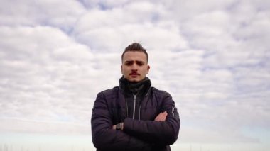 Ceketli genç adam beyaz bulutlara karşı kameraya poz veriyor.