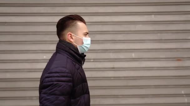 Facet w masce medycznej spaceruje po zamkniętych okiennicach sklepów. — Wideo stockowe