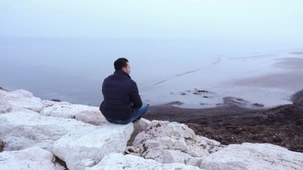 Mężczyzna w czarnej kurtce siedzi na dużych białych kamieniach w pobliżu zimnego morza — Wideo stockowe