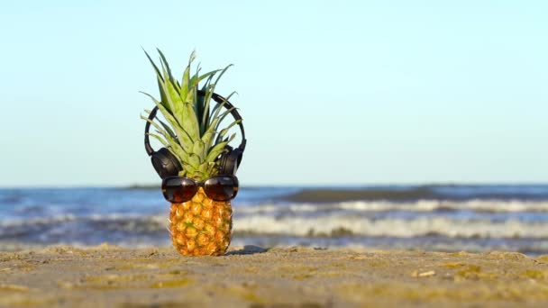 Pineapple with glasses and headphones on the sea sand — Stock Video