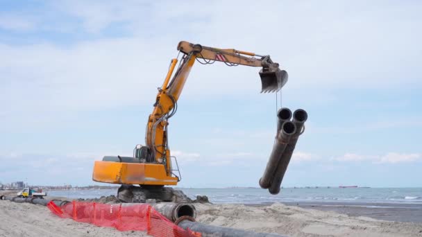 Orangefarbener Kettenbagger trägt drei Rohre aufs Meer — Stockvideo