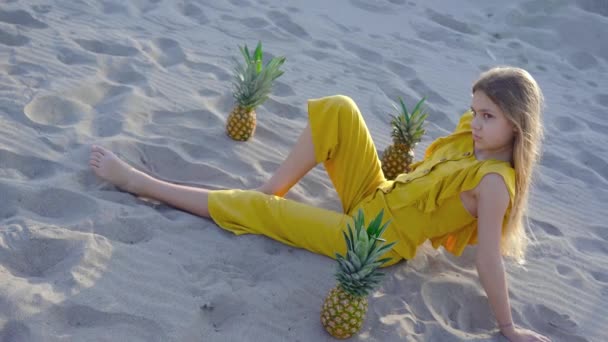 Giovane ragazza bionda seduta sulla sabbia tra gli ananas — Video Stock