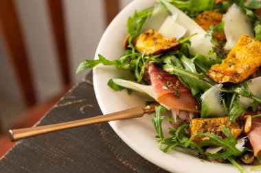 Hafif sonbahar salatası roka, fırında balkabağı, prosciutto ve taze parmesan, ahşap bir masada bir tabak salata.