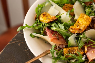 Hafif sonbahar salatası roka, fırında balkabağı, prosciutto ve taze parmesan, ahşap bir masada bir tabak salata.