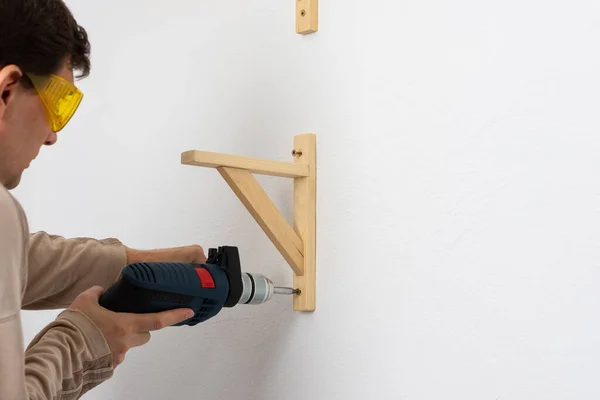 Bir adam beyaz bir duvardaki dairede bir raf asıyor, bir işçi bir rafın köşesinde matkapla vidaları sıkıyor.