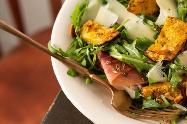 Hafif sonbahar salatası roka, fırında balkabağı, prosciutto ve taze parmesan, ahşap bir masada bir tabak salata.