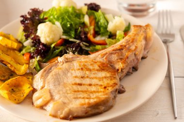 Kemiğin üzerinde kızarmış domuz eti, haşlanmış balkabağı parçaları ve yakın bir tabakta salata, karnabahar ve Napa lahana, çan biberi ve baharat ile yeşil salata.