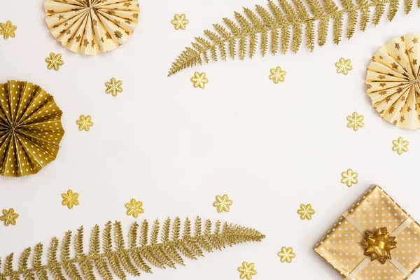 Festive white background with gold decoration , gift box and paper christmas tree decorations, glittering snowflakes and shiny golden fern leaves, top view, copy space