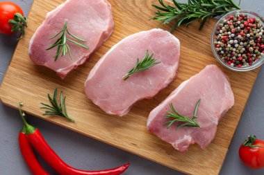 Baharatlı çiğ domuz eti, ahşap bir tahtada biberiye ve sebzeler, gri beton arka planda hazırlanmış et parçaları.