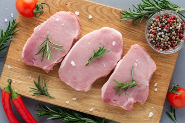 Baharatlı çiğ domuz eti, ahşap bir tahtada biberiye ve sebzeler, gri beton arka planda hazırlanmış et parçaları.