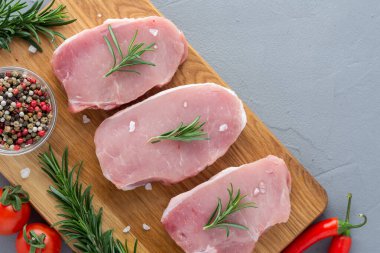 Baharatlı çiğ domuz eti, ahşap bir tahtada biberiye ve sebzeler, gri beton arka planda hazırlanmış et parçaları.