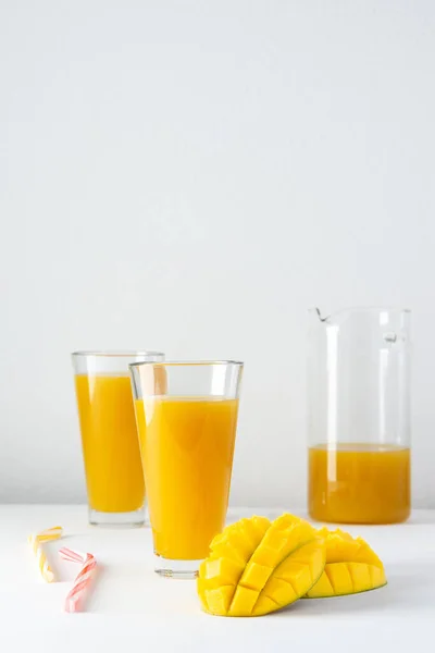 Mango juice in a glass jug and glasses with juice and a straw on a white background, chopped mango and whole, delicious healthy sweet natural drink