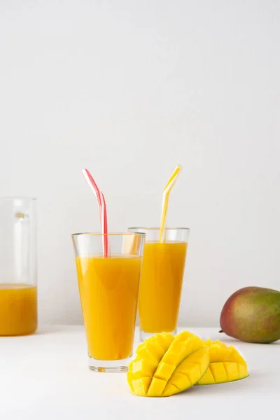 Mango juice in a glass jug and glasses with juice and a straw on a white background, chopped mango and whole, delicious healthy sweet natural drink