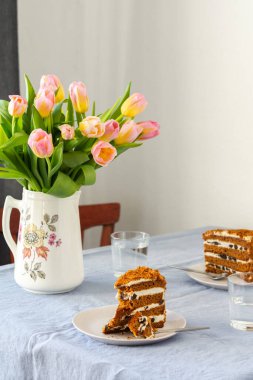 Bir sürahide pembe lale buketi, tabakta kuru erik ve mavi masa örtüsü olan bir masada su dolu porsiyonlar.