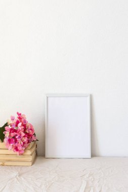 Ev dekorasyonu, beyaz boyanmış beton duvarın yanındaki boş resim çerçevesi, eski kitapların üzerindeki pembe ortanca dalı ve bir mum.