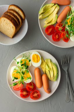 Lezzetli kahvaltı, sosisli haşlanmış yumurta, domates ve avokado, bezelye ve ayçiçeği mikro yeşillikleri, yemek tabakları ve gri arka planda bir kase ekmek, üst manzara.