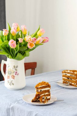 Bir sürahide pembe lale buketi ve masasında mavi bir masa örtüsü olan bir tabakta kuru erikli pasta.