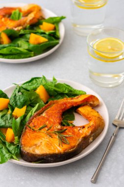 Ispanaklı kızarmış somon bifteği ve gri arka planda sarı domates, limonlu bir bardak su, sağlıklı kırmızı balıklarla doyurucu bir öğle yemeği.