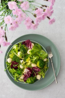 Masanın üst görüntüsü ve hafif salatalı bir tabak brokolili yeşil salata, mozzarella peyniri ve mikroyeşiller, vazoda pembe karanfiller.