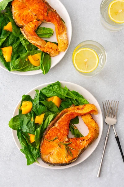Fried salmon steak with spinach and yellow tomatoes in a plate on a gray background, a glass of water with lemon, a hearty lunch with healthy red fish