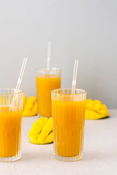 Freshly squeezed mango juice in glasses with a straw, ripe cut mango on a gray background