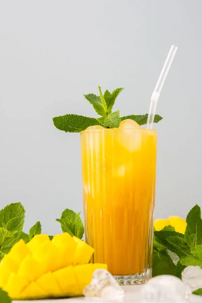 Freshly squeezed mango juice with ice in glass with a straw, ripe cut mango with mint on a gray background