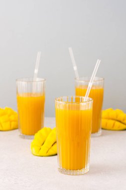 Taze sıkılmış mango suyu bardakta pipetle, olgun kesilmiş mangoyla gri arka planda.