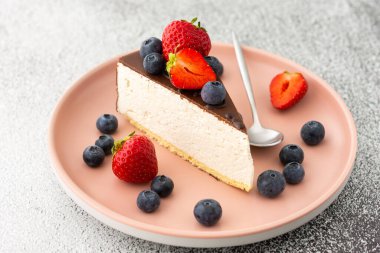 Çikolatayla kaplı bir parça cheesecake. Yaban mersinli ve çilekli. Tatlı kaşığıyla pembe bir tabakta yakın plan.