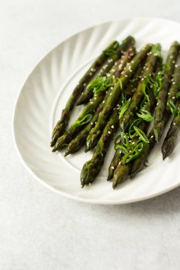 Taze otlarla kaplı buharda pişirilmiş kuşkonmaz, beyaz bir tabakta güzelce sıralanır. Yemek canlı görünüyor ve sağlıklı bir yemek ya da yan yemek için uygun.