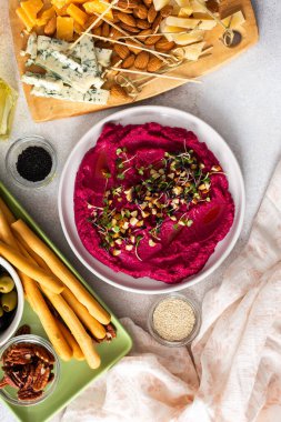 Beetroot hummus is placed in the center of a charcuterie board, surrounded by snacks like olives, nuts, cheese, and crispy breadsticks. A light, inviting setup showcases vibrant colors