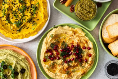 A variety of hummus flavors including turmeric, classic, and broccoli are served with fresh bread, nuts, and garnishes on a colorful charcuterie board, perfect for sharing.