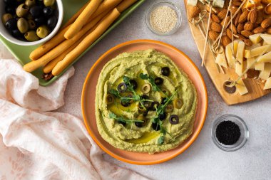 Broccoli hummus is artistically displayed in a bowl, garnished with olives. Accompanied by a charcuterie board featuring cheese, nuts, and breadsticks for a delightful snack experience.