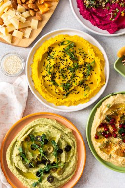 A variety of hummus flavors including turmeric, classic, and broccoli are served with fresh bread, nuts, and garnishes on a colorful charcuterie board, perfect for sharing