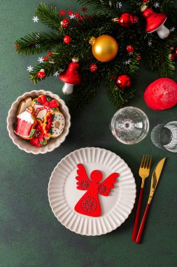 İçinde boş bir tabak, peçete ve zencefilli ekmek, çatal bıçak takımı ve koyu yeşil arka planda bir mum olan bir Noel masası. Bahar dalları ve kırmızı ağaç süslemeleri şenlikli bir atmosfer yaratır.