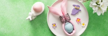 A beautifully arranged table setting for Easter features a pink napkin wrapped around an egg, surrounded by floral decorations and colorful candies, banner