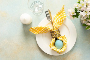 A beautifully arranged Easter table setting showcases a decorative egg nestled in a yellow napkin, surrounded by a glass of water and fresh flowers, perfect for holiday gatherings.