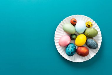 Paskalya yumurtalarının çeşitli renkleri ve desenleri yuvarlak beyaz bir tabakta parlak mavi bir arka planda sergilenir. Bayram kutlamaları için mükemmeldir.