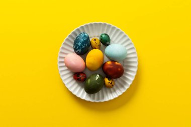 A variety of beautifully painted Easter eggs in different colors and patterns are placed on a delicate white plate. The vibrant hues stand out against the cheerful yellow backdrop