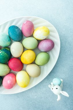 Mavi arka planda dekoratif tavşanları olan oval bir tabakta çok renkli Paskalya yumurtaları, üst manzara, mutlu Paskalya tatili